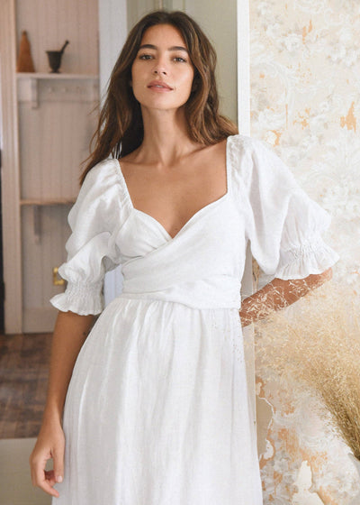 White linen dress with a soft v neckline, wrap tie, tiered skirt, and puffed sleeves.