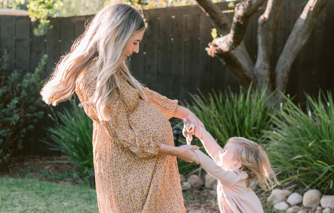 Pregnant woman glowing in soft floral maxi maternity dress by LIV & Co, joyfully playing with her daughter.